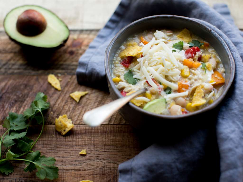 Aerial shot of a bowl of light chili with vegetables and cheese on top next to an avocado on a wooden tabletop.