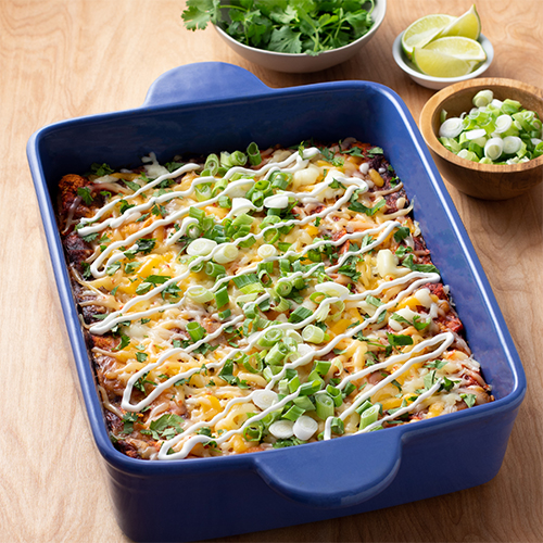 Enchiladas topped with cheese, green onions, and sour cream in a blue casserole. Limes, cilantro, and green onions in side dishes behind.