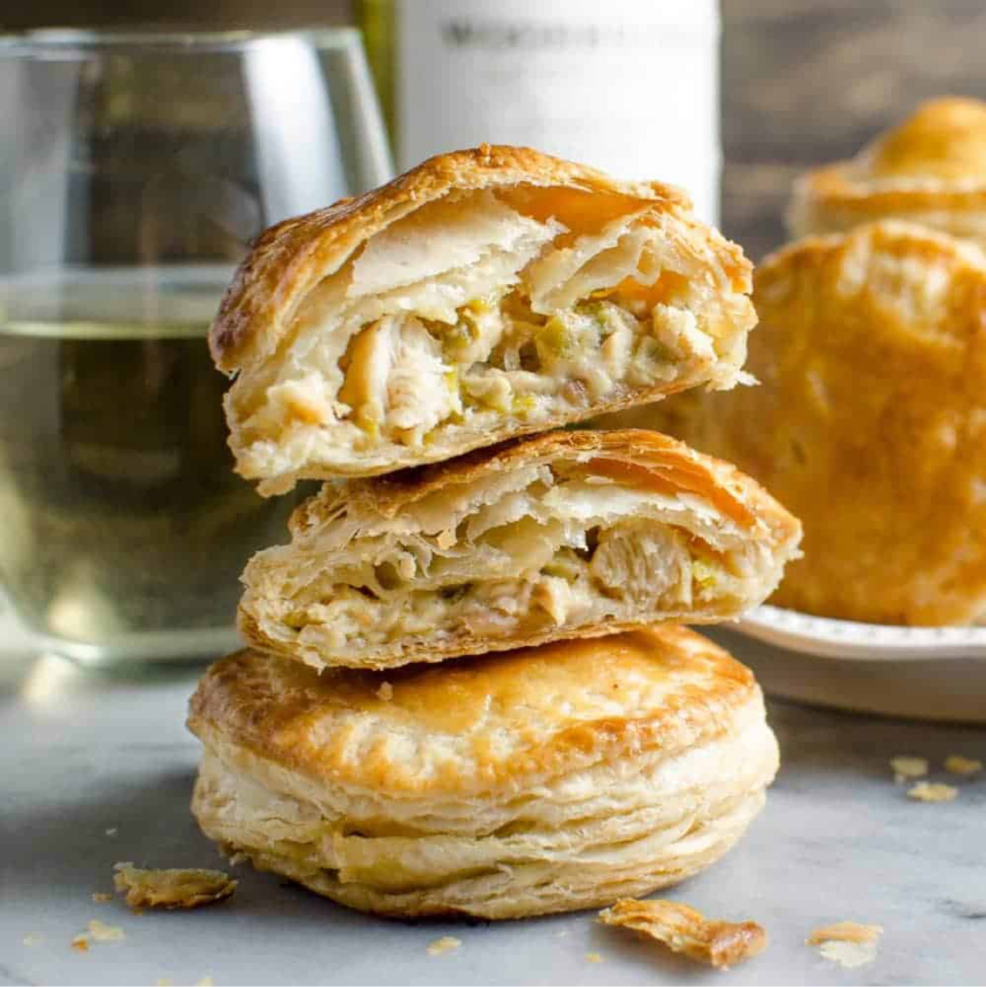 Three cheddar chicken and vegetable pies sticked on top of each other. Glass of white wine and more pies in the background.