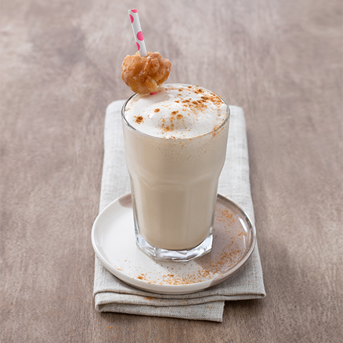 Coffee and donuts smoothie in a clear glass on top of a small cream plate and grey napkin. A pink polka dot straw with a donut hole and cinnamon dusted on top of the smoothie.