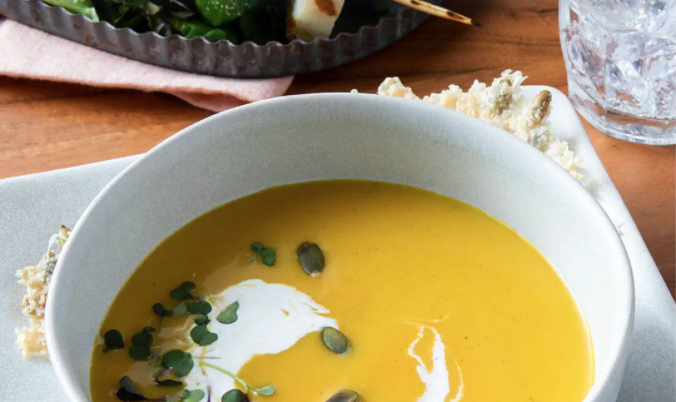 Butternut Squash Soup in a white bowl with pepitas and microgreens on top. Pepita crisps on a white plate on the side.