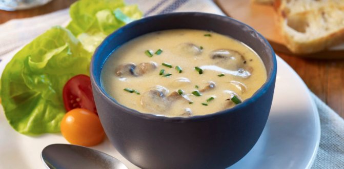 Mushroom cheddar soup in a black bowl, on a white plate with lettuce and tomatoes on the side and a silver spoon.
