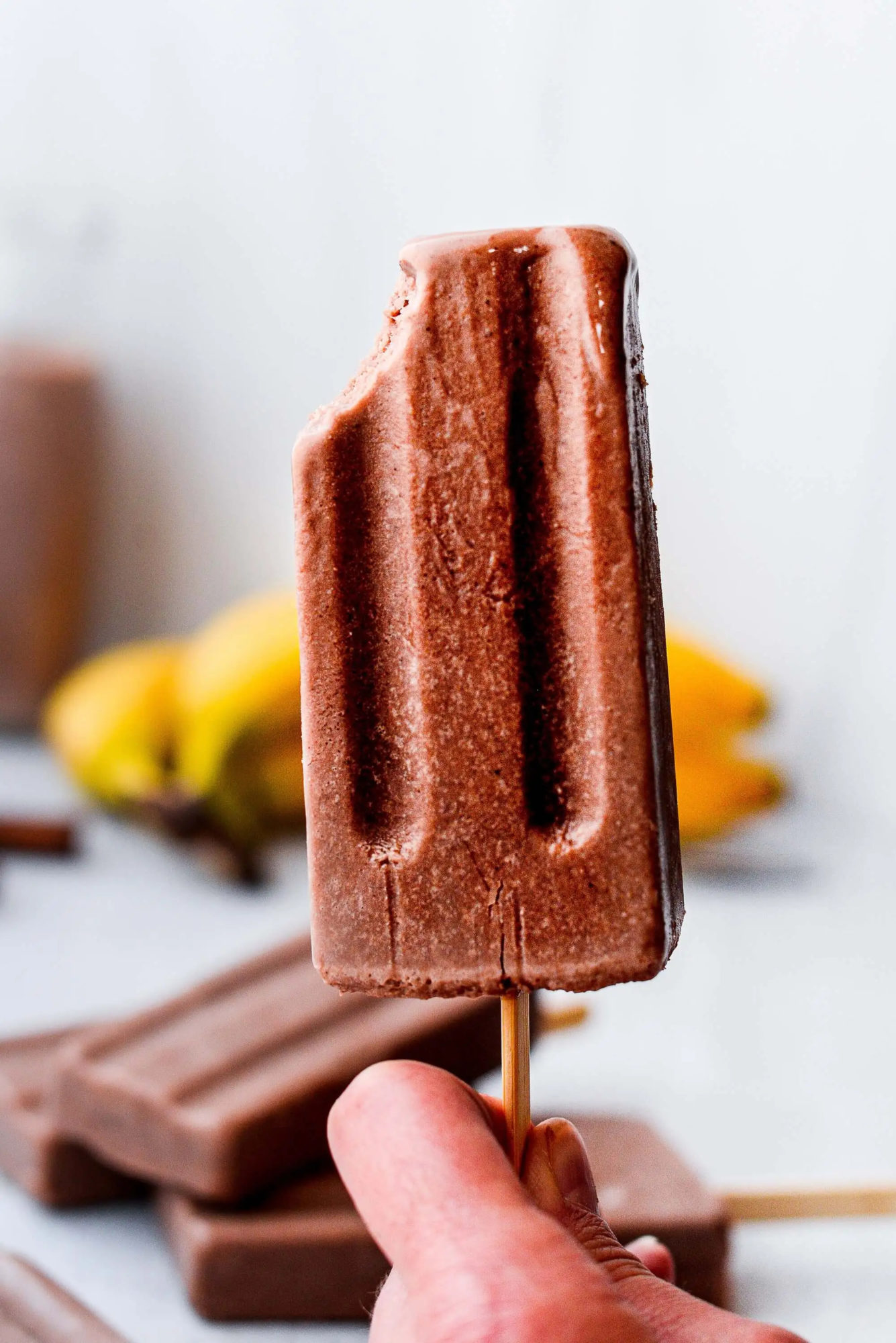 A hand holding a Chocolate Fudgesicle, with bananas, chocolate, and more fudgesicles blurred in the background.