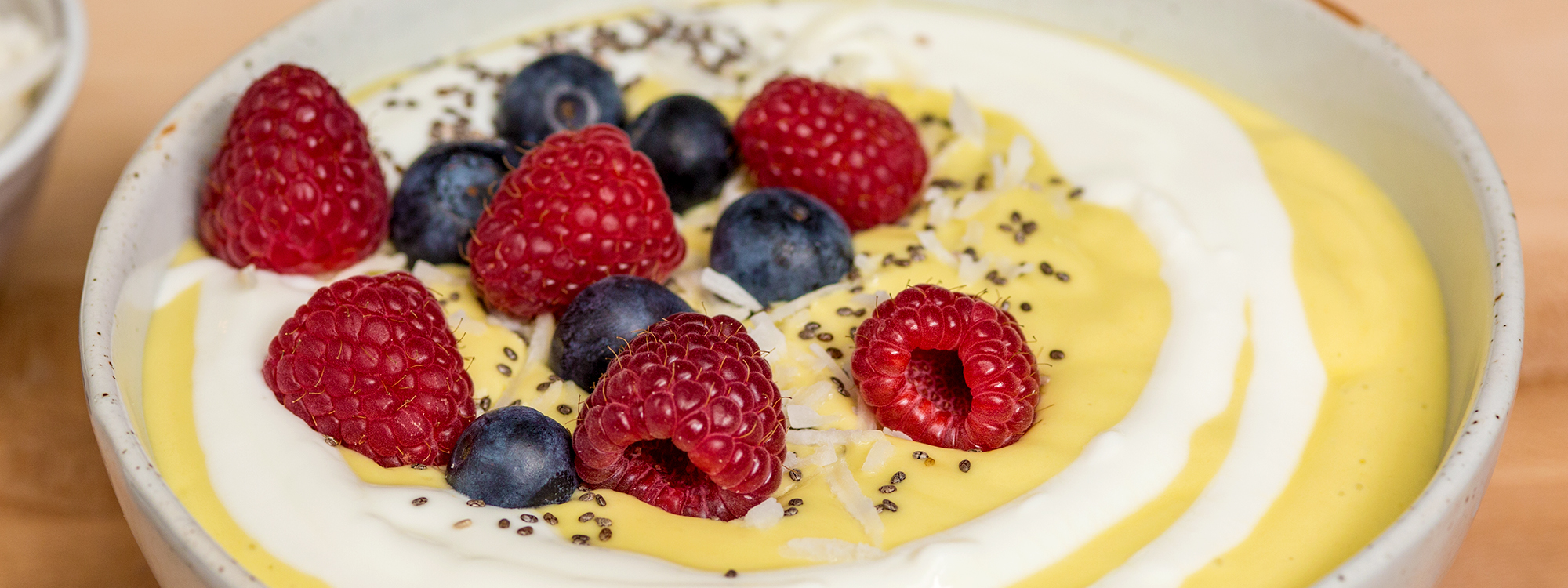 Mango Avocado smoothie bowl with yogurt drizzle, raspberries, blueberries, coconut, and chia seeds on top. In a white bowl.