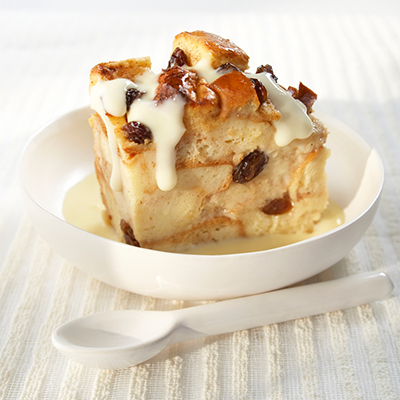 Cinnamon Raising Bread Pudding with Vanilla Sauce drizzled on top and pooling in a white bowl. White spoon on the side.