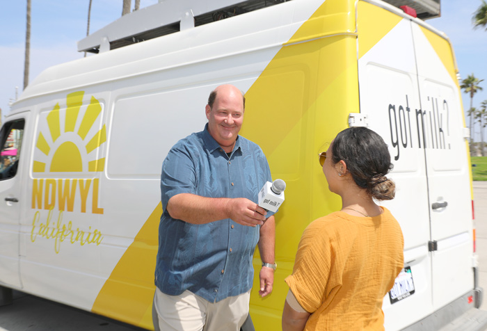 Image of Brian Baumgartner interviewing a woman, holding a microphone. standing in front of a news van