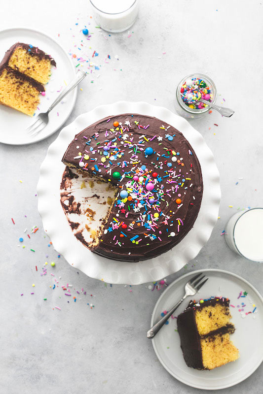 Aerial shot of a yellow cake with chocolate frosting and rainbow of sprinkles. On a white cake platter, two slices of cake on white plates, and with a glass of milk