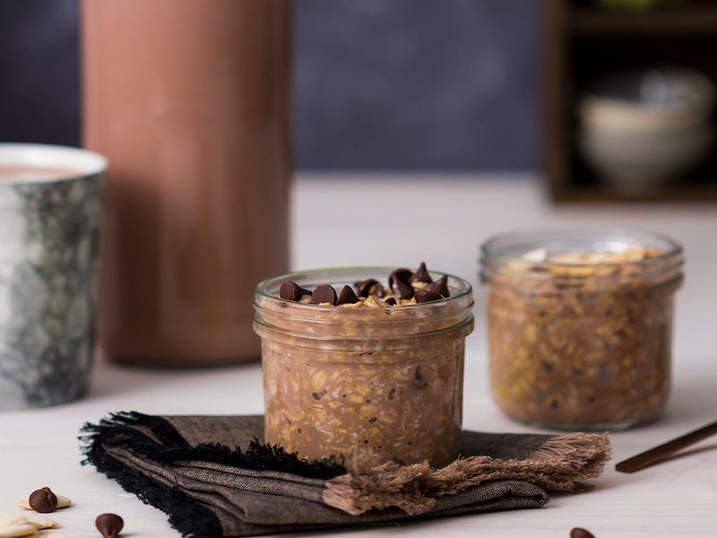 Two mason jars of chocolate overnight oats, with chocolate chips on top. Dishes in the background.