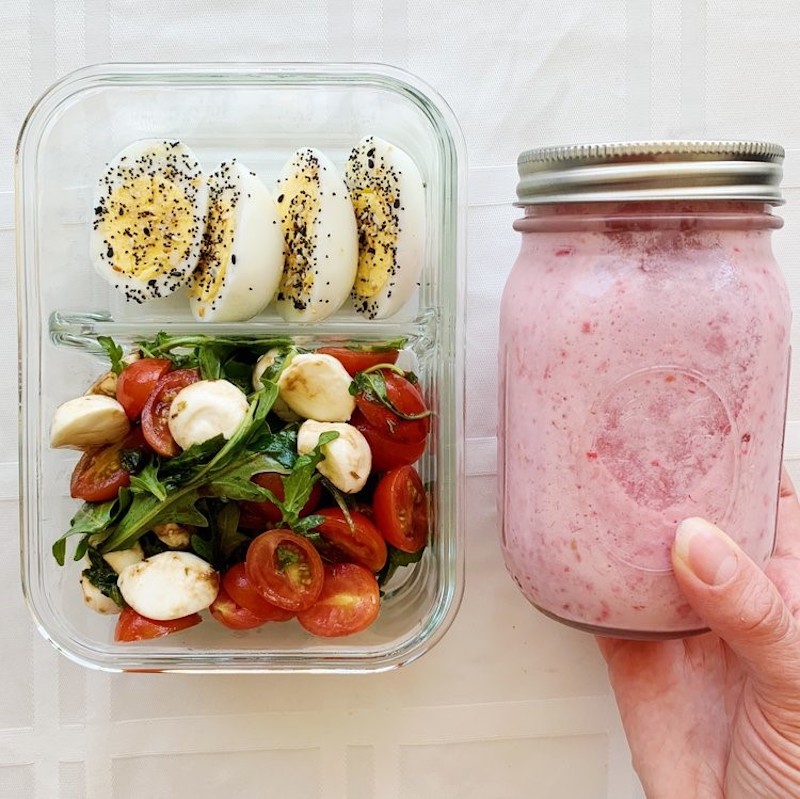 Harboiled eggs cut in half in a lunch box with an arugula salad. Pink strawberry smoothie in a mason jar held next to it.