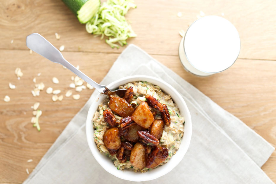 aerial shot of a bowl of zoats with caramelized pears and pecans on top. glass of milk and shredded zucchini/ oats on the table.