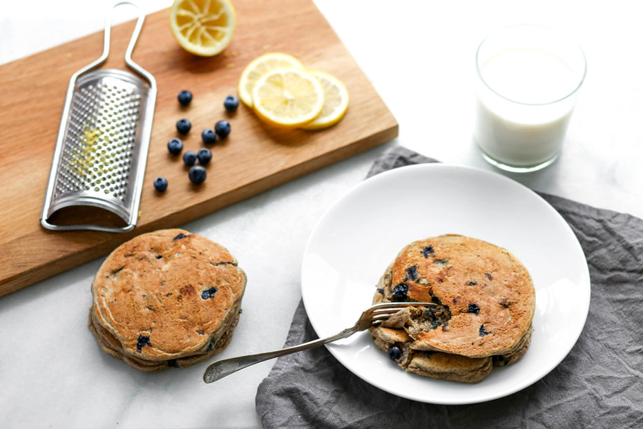 aerial shot of blueberry pancakes on a plate, cutting board with lemon and blueberry and a glass of milk in the background.