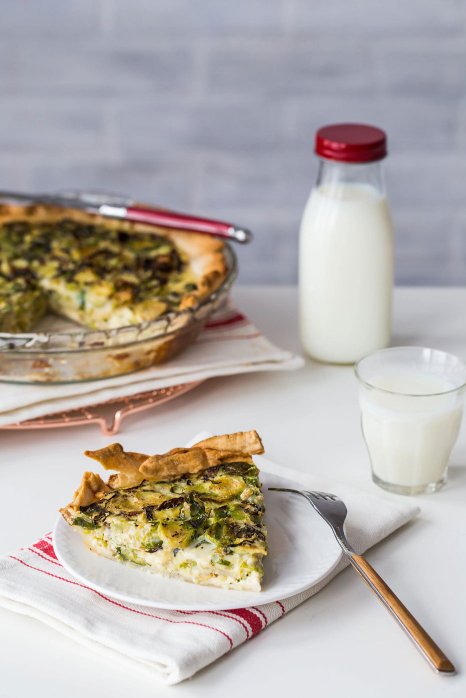 piece of brussel sprout quiche on a plate with a glass of milk, bottle of milk, and the rest of the quiche in the background.