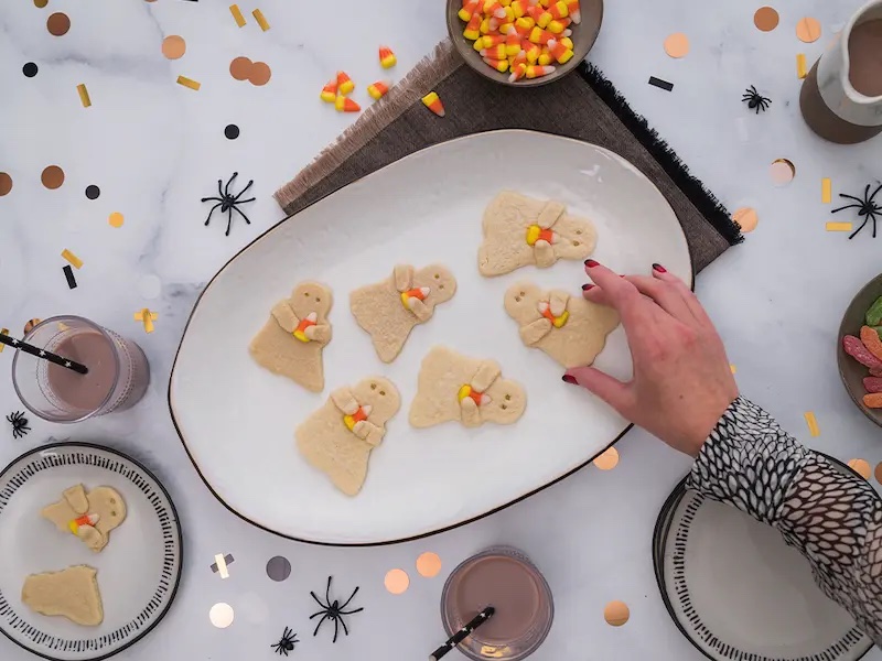 aerial shot of a plate of ghost halloween cookies. spider and halloween decor on table, with a glass of chocolate milk