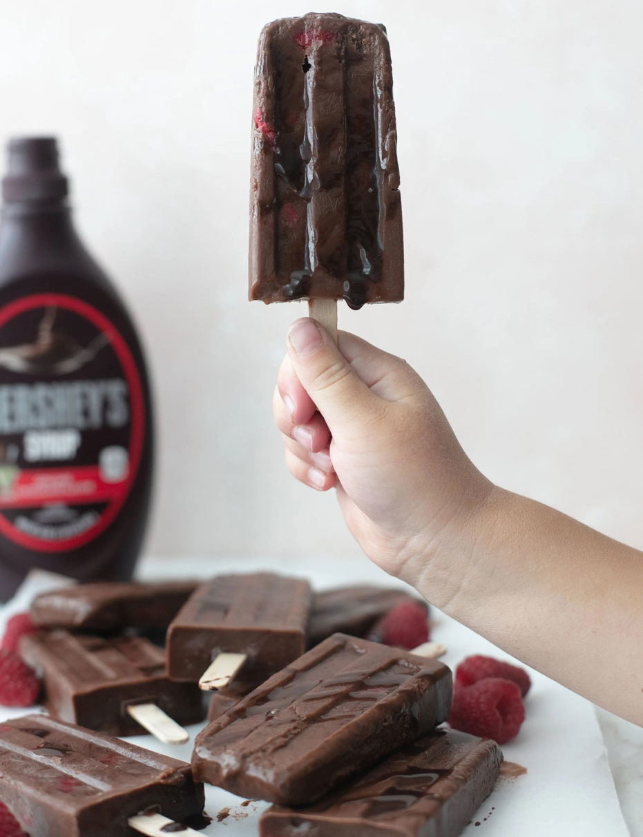 child's hand holding frozen chocolate bar on a stick with Hershey's syrup in the background