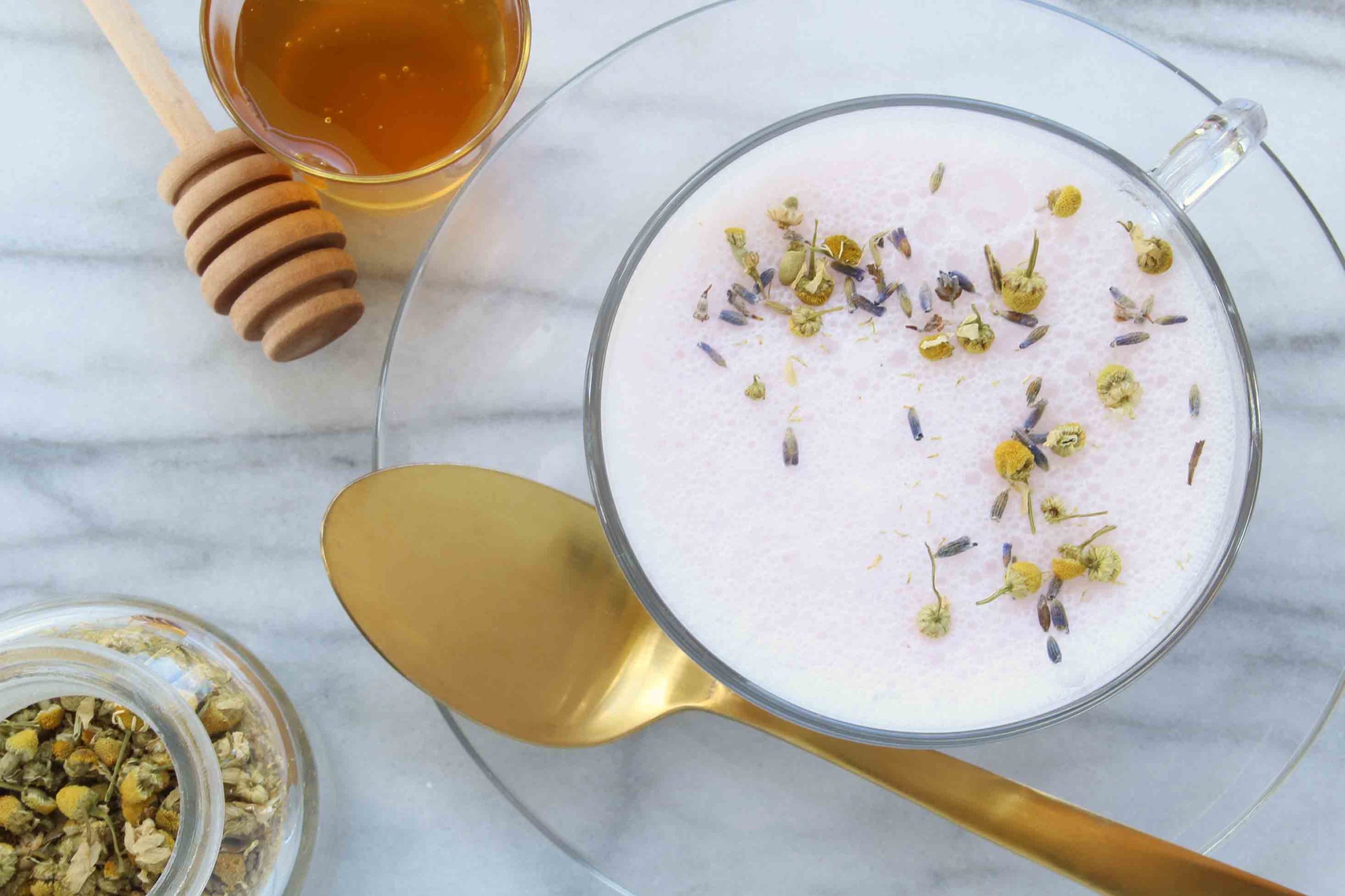 purple foamy drink with loose lavendar on top inn clear glass with honey and gold spoon on the table