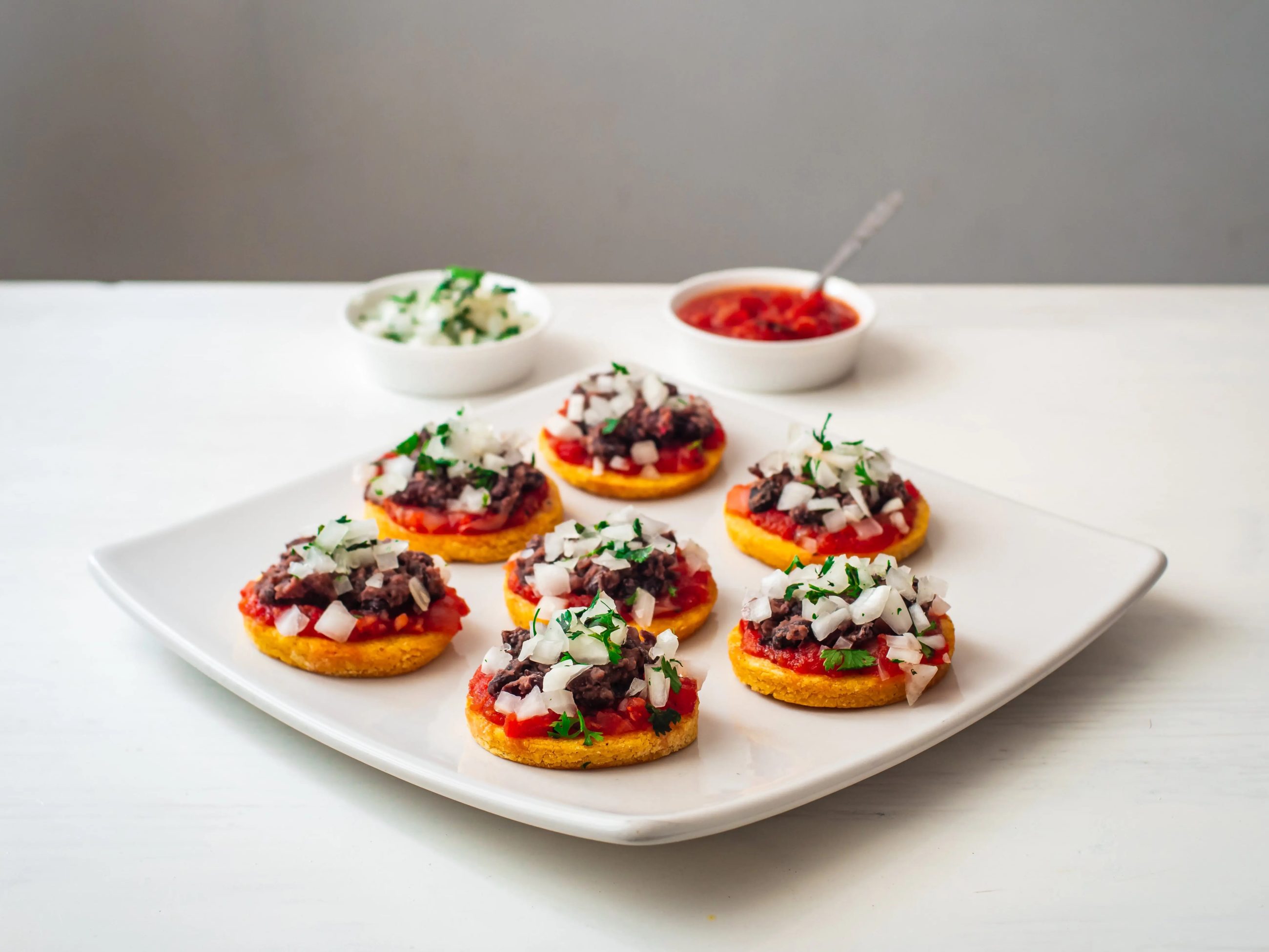 small bite size sopes with red suace, onions, beans on a white plate and white table.